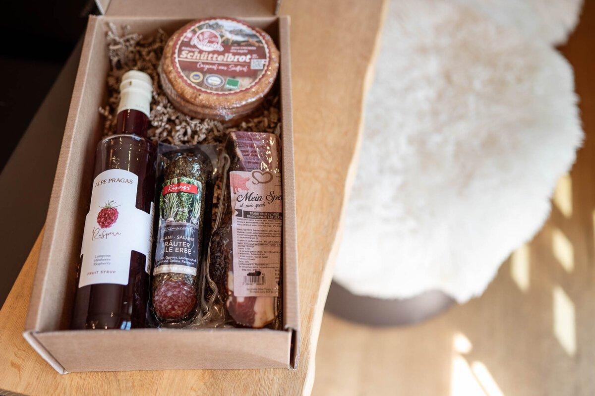 A gift box with various food items. Inside, there is a bottle of vinegar, a sausage, baked goods, and another treat.