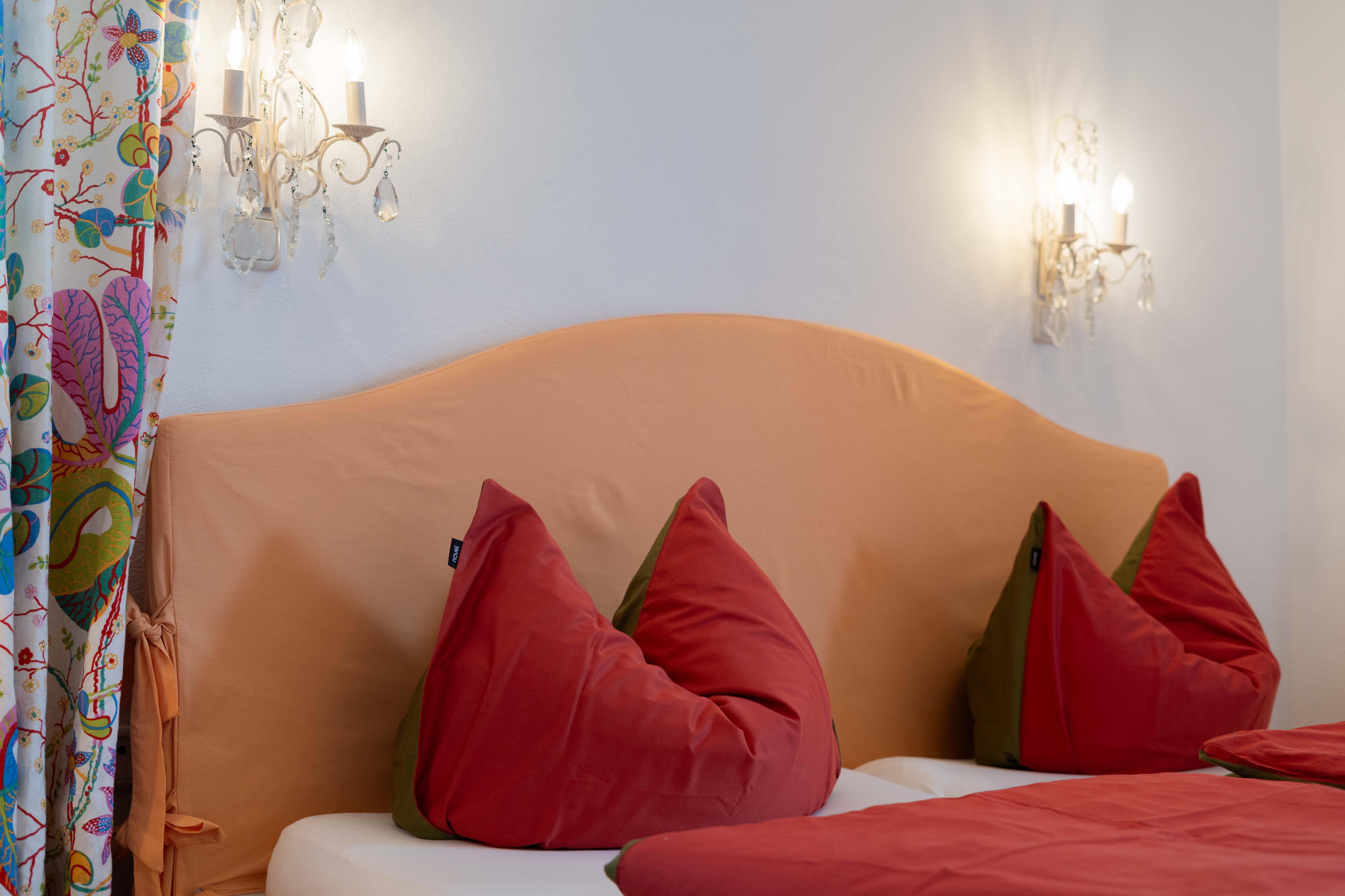 A cozy bedroom with a large headboard and red pillows. The walls are light, and there are decorative wall lights.