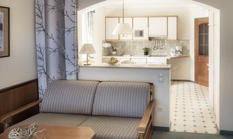 A cozy living room with a sofa and a wooden coffee table. In the background, there is a modern kitchen with white cabinets and a friendly atmosphere.