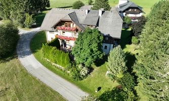 A large, charming house with a shingled roof, surrounded by trees and a well-maintained garden. A beautiful landscape with green meadows in the background.