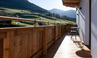 A balcony with a wooden railing and two chairs. In the background, green hills and mountains stretch under a clear sky.
