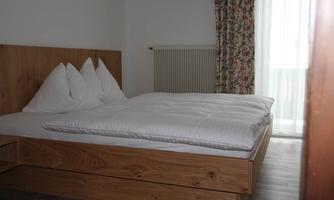 A simple bedroom with a large double bed and white pillows. The room has light walls and a curtain with a floral pattern.