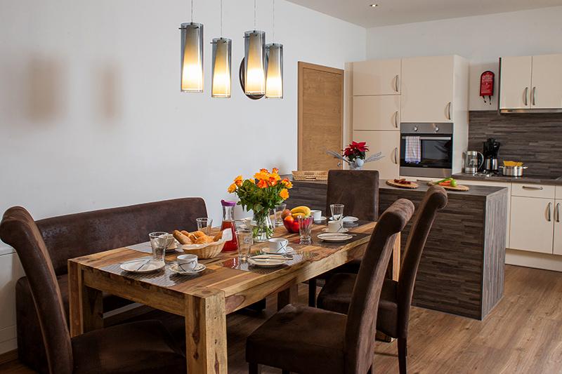 An inviting dining area with a large wooden table decorated with flowers and tableware. In the background, there is a modern kitchen with bright shelves and appliances.