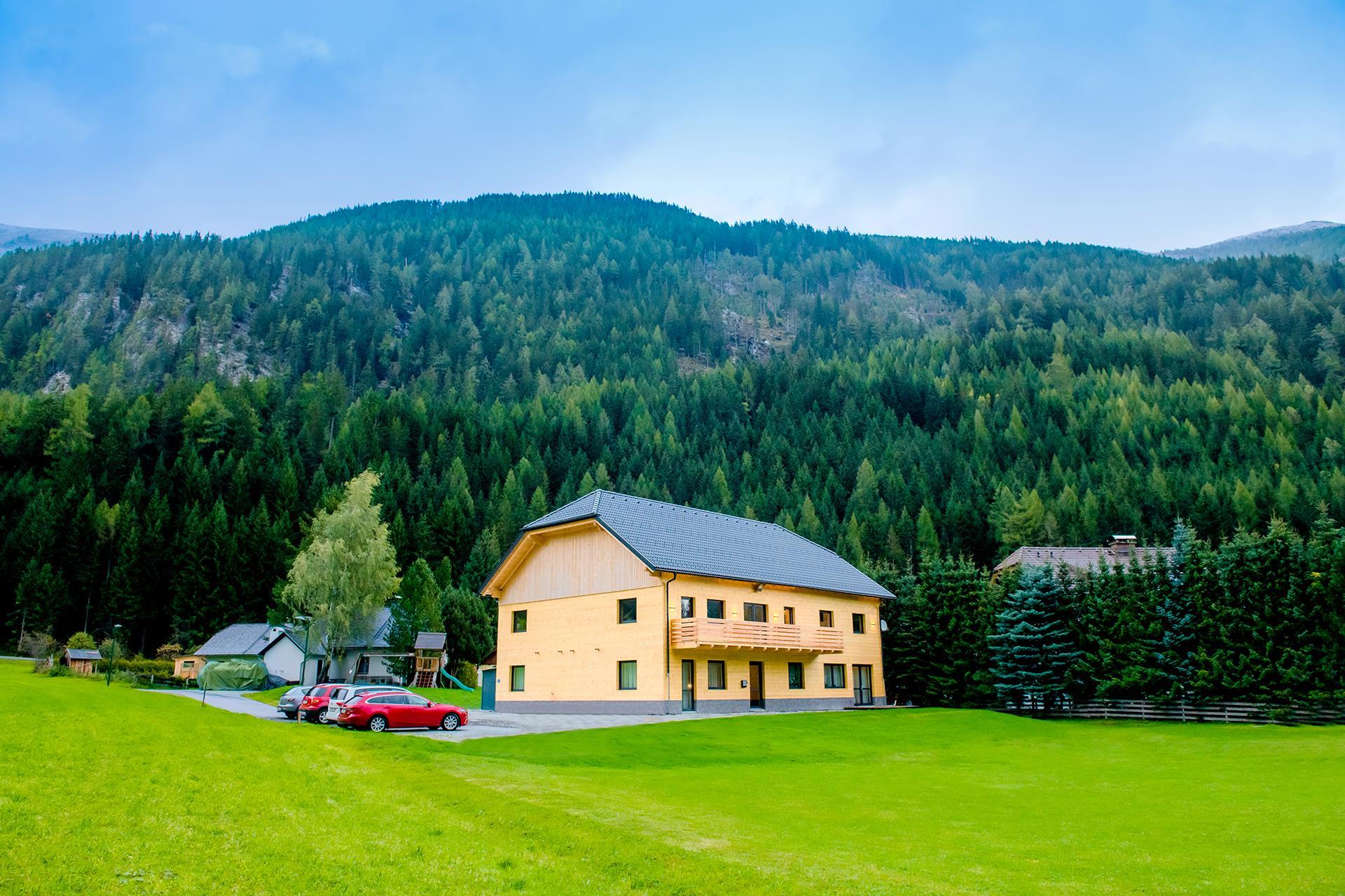 A yellow building in a green landscape, surrounded by trees and mountains. There are some cars on the property, and the sky is cloudy.