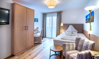 A bright room with a bed, a desk, and a comfortable armchair. Large windows provide daylight and a cozy atmosphere.