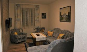 A cozy living room with blue sofas and a wooden table. Pictures hang on the walls, and the window is decorated with curtains.