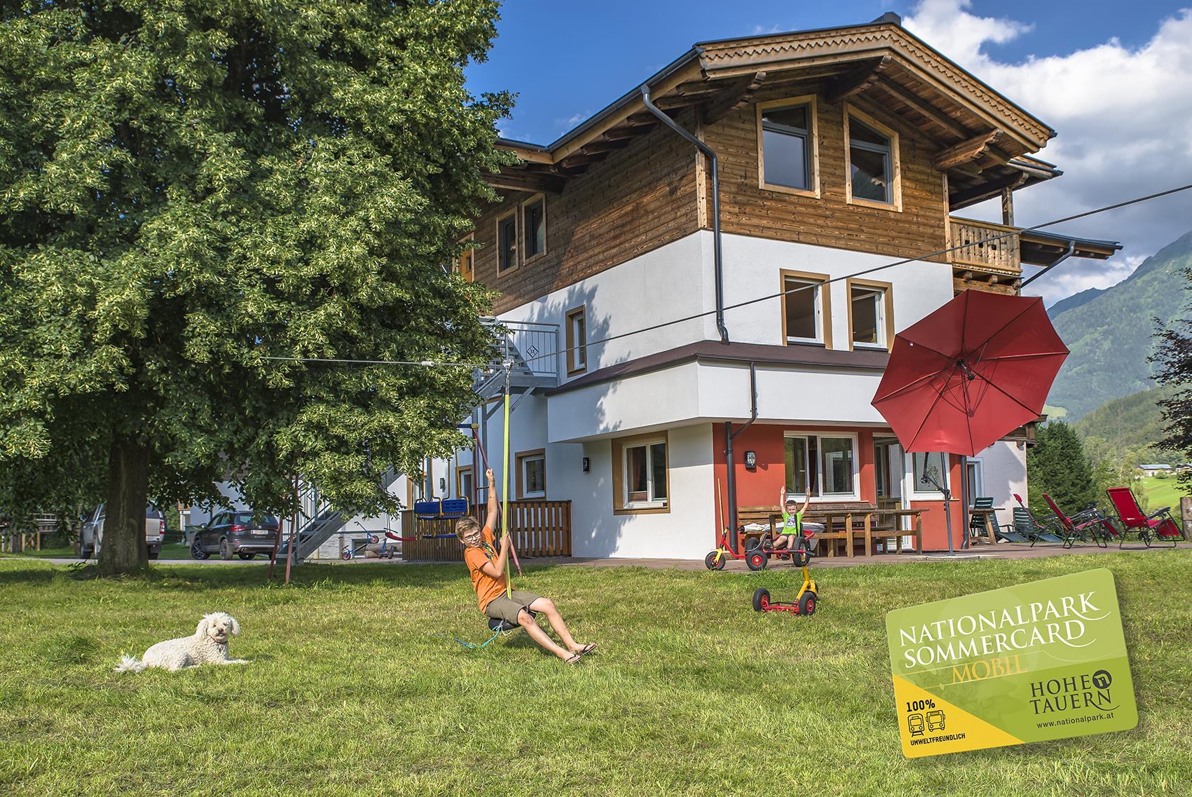 A beautiful house with a large garden, where a child is sitting on a swing. A dog is lying relaxed on the grass, and a parasol provides shade.
