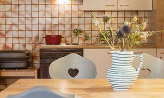 A cozy kitchen with a wooden table and chairs with heart-shaped cutouts. On the table, there is a vase with fresh flowers.