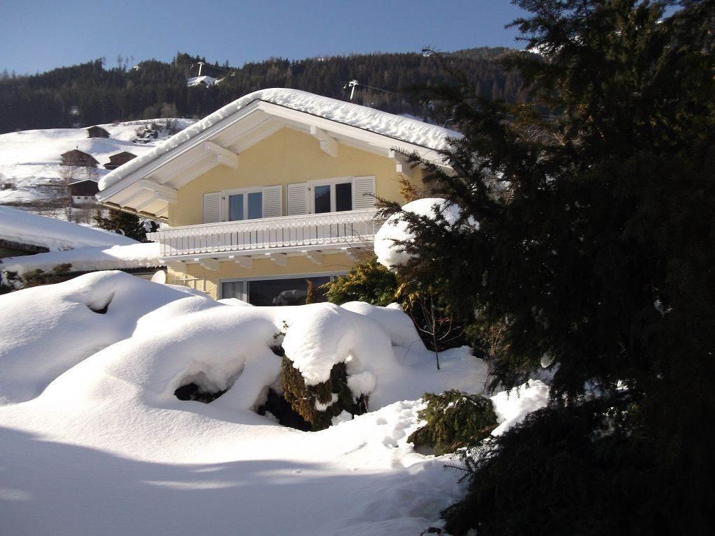 A cozy house in the snow, surrounded by snow-covered hills. The sun is shining on the wintry landscape.