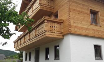 A modern wooden house with two balconies and a white facade. In front of the house, there is a bench and a bicycle.