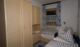 A cozy bedroom with two beds and a large wooden wardrobe. The windows are adorned with curtains and there is a warm, inviting atmosphere.