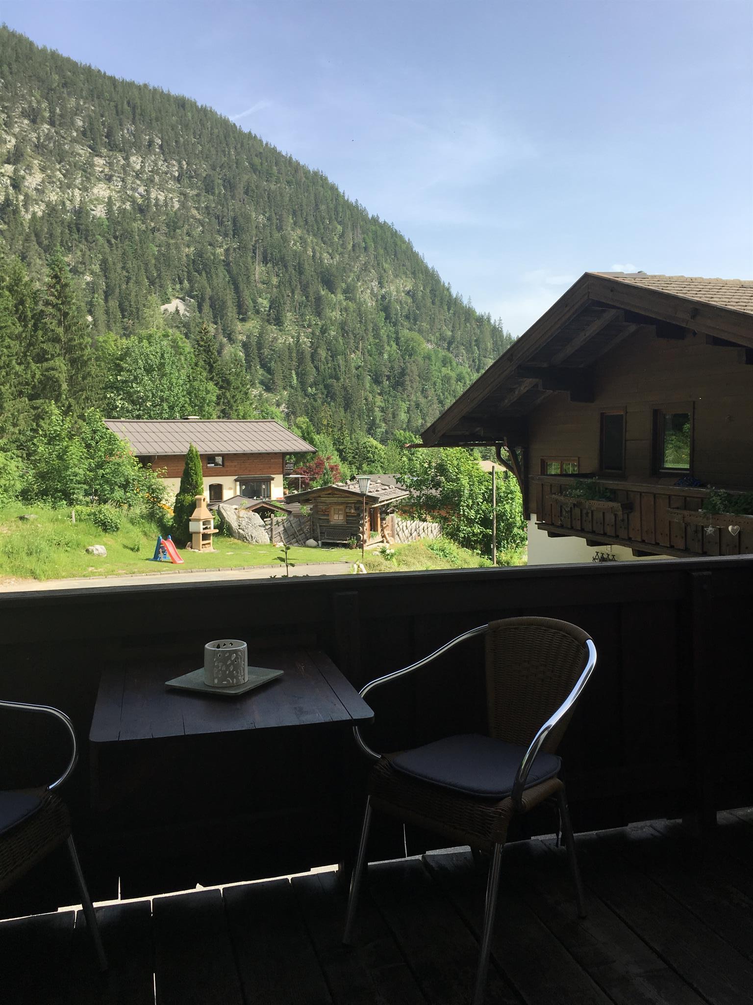 A cozy balcony with chairs and a small table. In the background, mountains and traditional wooden houses are visible.
