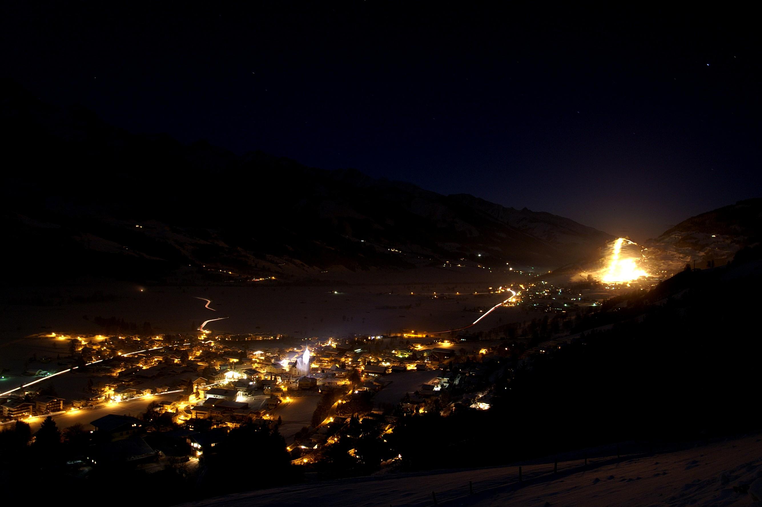 A picturesque mountain town at night, illuminated by warm lights. The surrounding landscape is dark, making the town stand out particularly.