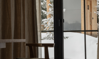 A cozy room with a wooden chair and a large window front. Outside, there is a wooden house surrounded by snow-covered trees.