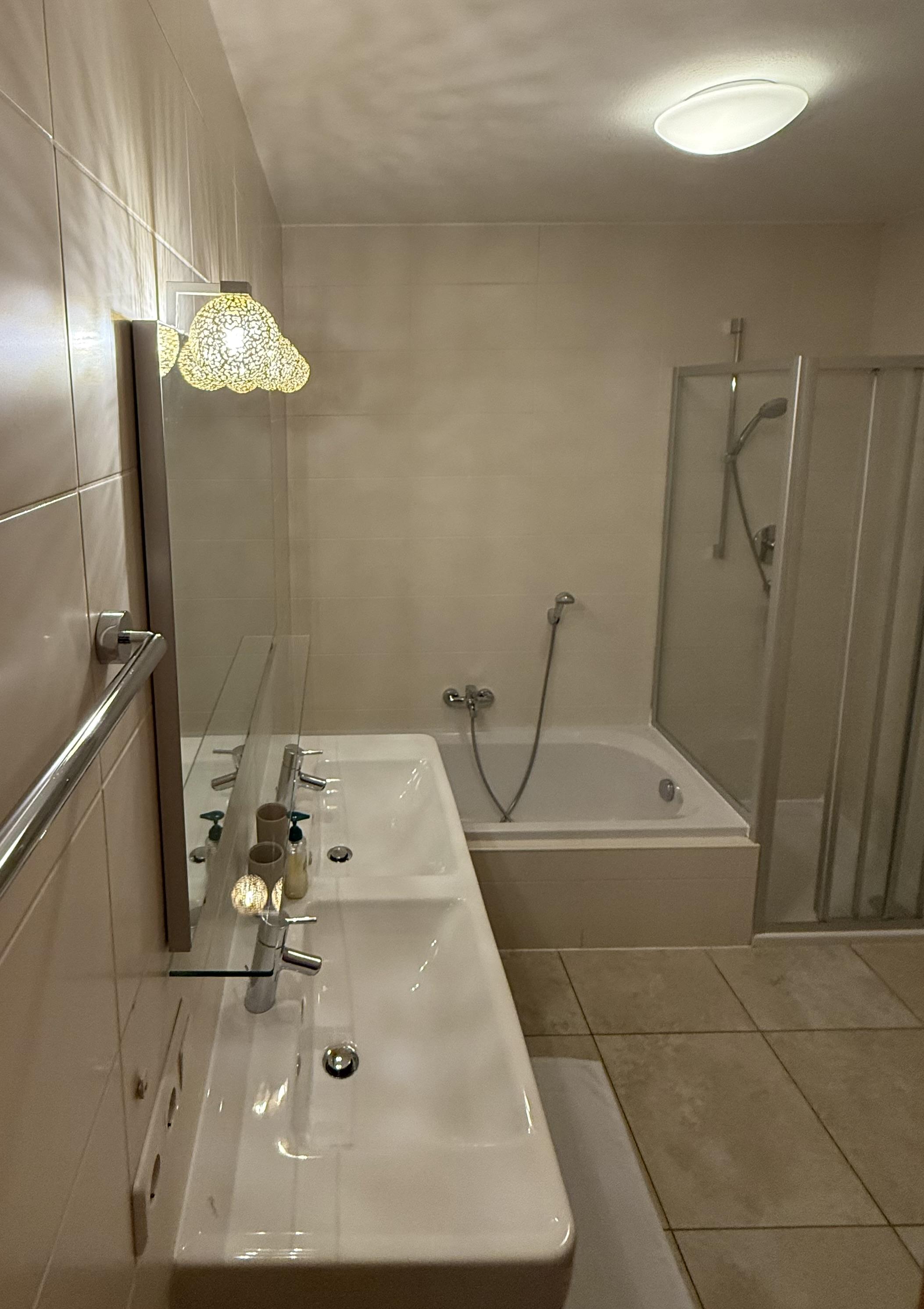 A modern bathroom with two sinks, a bathtub, and a separate shower. The walls are light tiled, and there is an elegant light source.