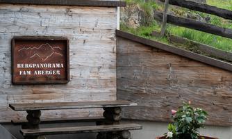 A wooden sign with the inscription "Mountain Panorama Fam Aberger" hangs on a rustic wall. In front, there is a bench and a flower pot with a plant.