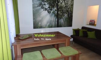 A cozy living room with a wooden table and green seating cushions. A large picture of a forest with sunlight rays hangs on the wall.