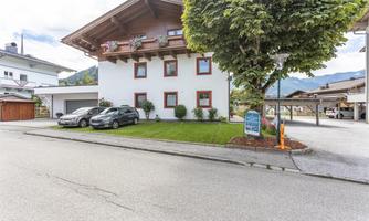 A bright building in alpine style with red shutters. In front of the house are two cars and a well-kept lawn.