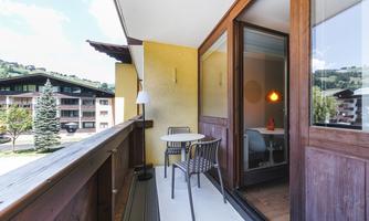 A balcony with two chairs and a small table. In the background, green mountains and a quiet residential complex are visible.