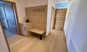 A bright hallway with a wooden floor and light walls. On the left side, there is a wardrobe with a seating area and several hooks.