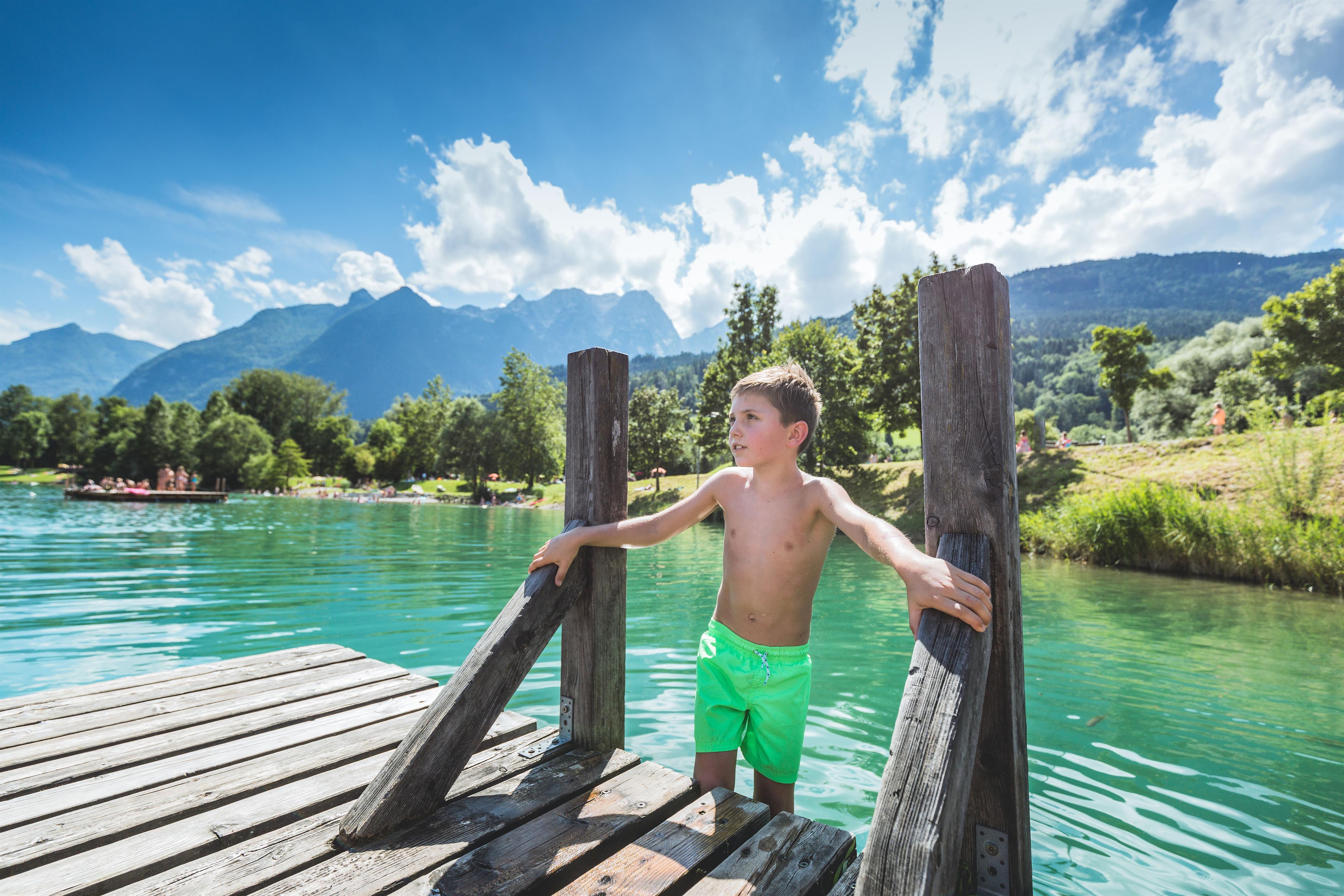 A boy stands on a dock and looks at the clear water. In the background, mountains and trees can be seen, all under a blue sky.