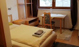 A simple room with a single bed, a table, and a chair. Large windows let in a lot of light and offer a view outside.