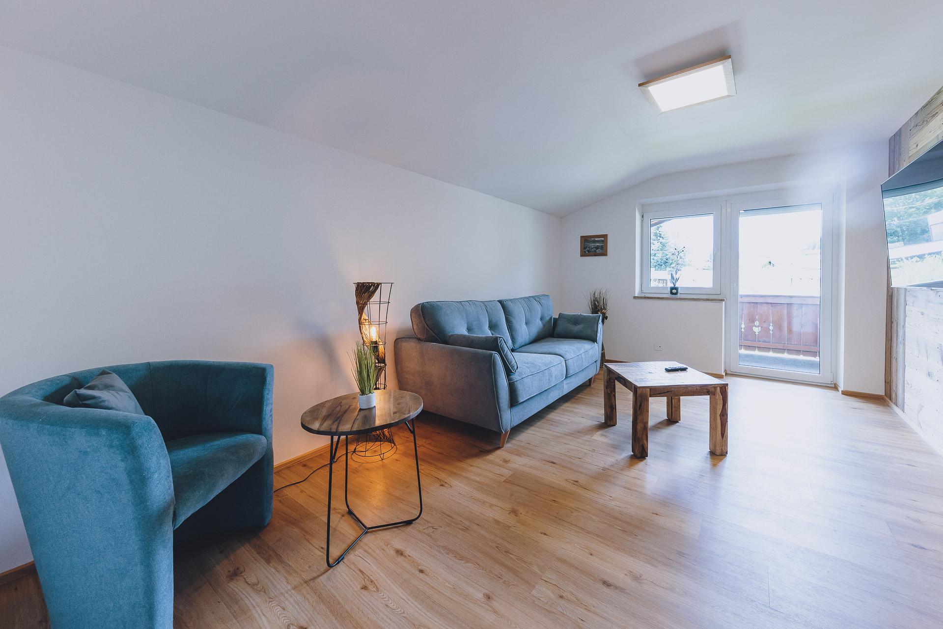 A modern living room with a blue sofa, an armchair, and a wooden table. Large windows let in plenty of light and provide access to the balcony.