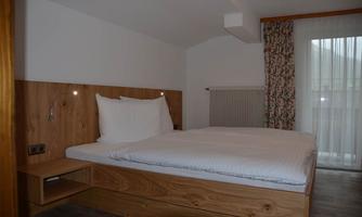 A simple bedroom with a wooden bed and white bedding. Windows with curtains let in plenty of light.
