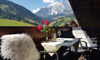 A beautiful balcony with a view of the mountains and a green landscape. On the table are wine and fresh flowers.