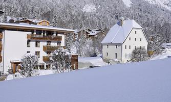 A snowy landscape with traditional alpine houses and mountains in the background. The snow covers the roofs and the ground, creating a peaceful winter atmosphere.
