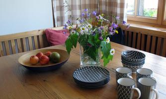A table with a bowl full of apples and a vase with colorful flowers. On it are plates and coffee cups in various designs.