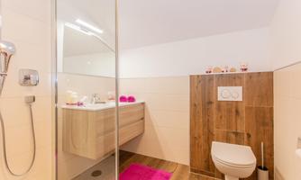 A modern bathroom with a spacious shower and a wood element. The colors are bright and inviting, with a pink rug and cosmetic items on the sink.