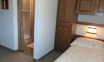 A simple bedroom with a bed and wooden cabinets. In the background, a bathroom with a shower can be seen.