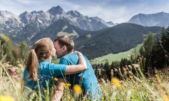 A couple is sitting in the grass and embracing each other in a picturesque mountain landscape. In the background, there are tall mountains and green meadows.