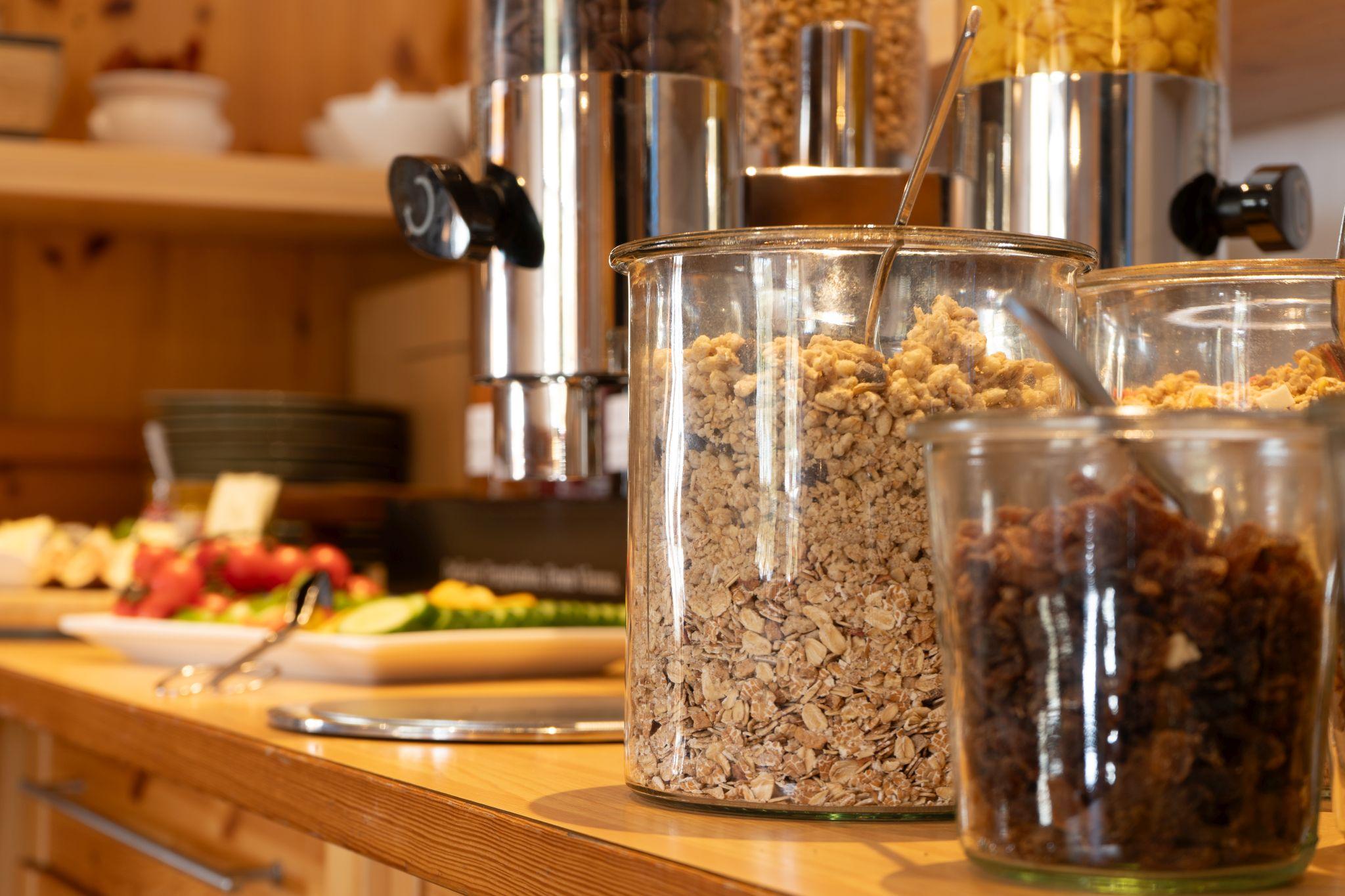 A buffet with various types of muesli in glass containers. Fresh fruit and dishes are visible in the background.