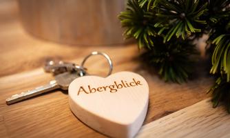 A heart-shaped wooden keychain with the inscription "Abergblick" is lying on a wooden table. Next to it are some green fir branches.