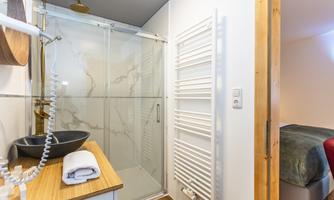 A modern bathroom with a glass shower and an elegant sink. In the background, a towel warmer and a bedding area are visible.
