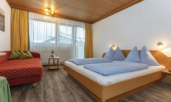 A cozy room with two beds and blue pillows. A red sofa and large windows create a bright, inviting atmosphere.