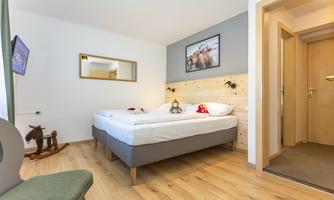A cozy hotel room with two beds and a wooden floor. A mirror and a picture hang on the wall, and a small chair stands next to it.