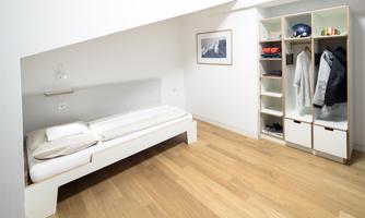 A bright bedroom with a single bed and a wardrobe. The room has a modern, minimalist style and natural wooden floors.