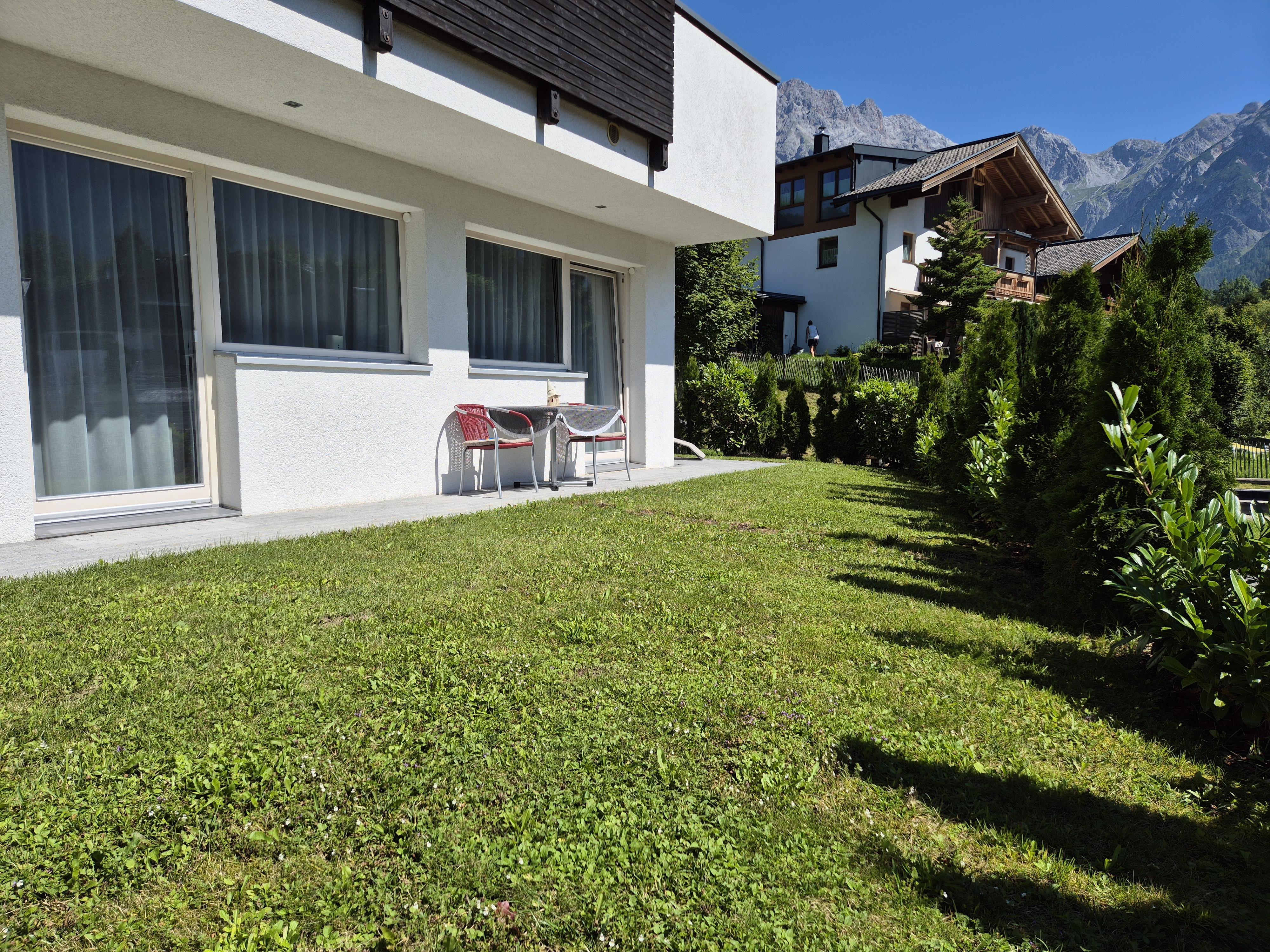 A modern building with large windows and a small seating area outside. In the background, green hills and mountains are visible.