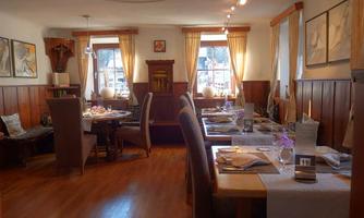 A cozy restaurant with wooden walls and large windows. Tables are set for guests, and artworks decorate the walls.