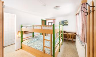 A bright room with a bunk bed and simple furniture. There is a desk and an armchair next to a window.