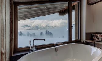 A modern bathroom with a freestanding bathtub and a large window. The view shows snow-covered mountains and a misty landscape.