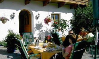 A cozy terrace with several people enjoying coffee and pastries. Surrounded by blooming plants and an inviting home.