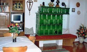 A cozy living space with a large, green tiled stove. The table is set and flowers in the background create a friendly atmosphere.
