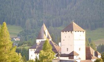 A picturesque castle with towers and impressive architecture. In the background, green mountains and forests stretch out.