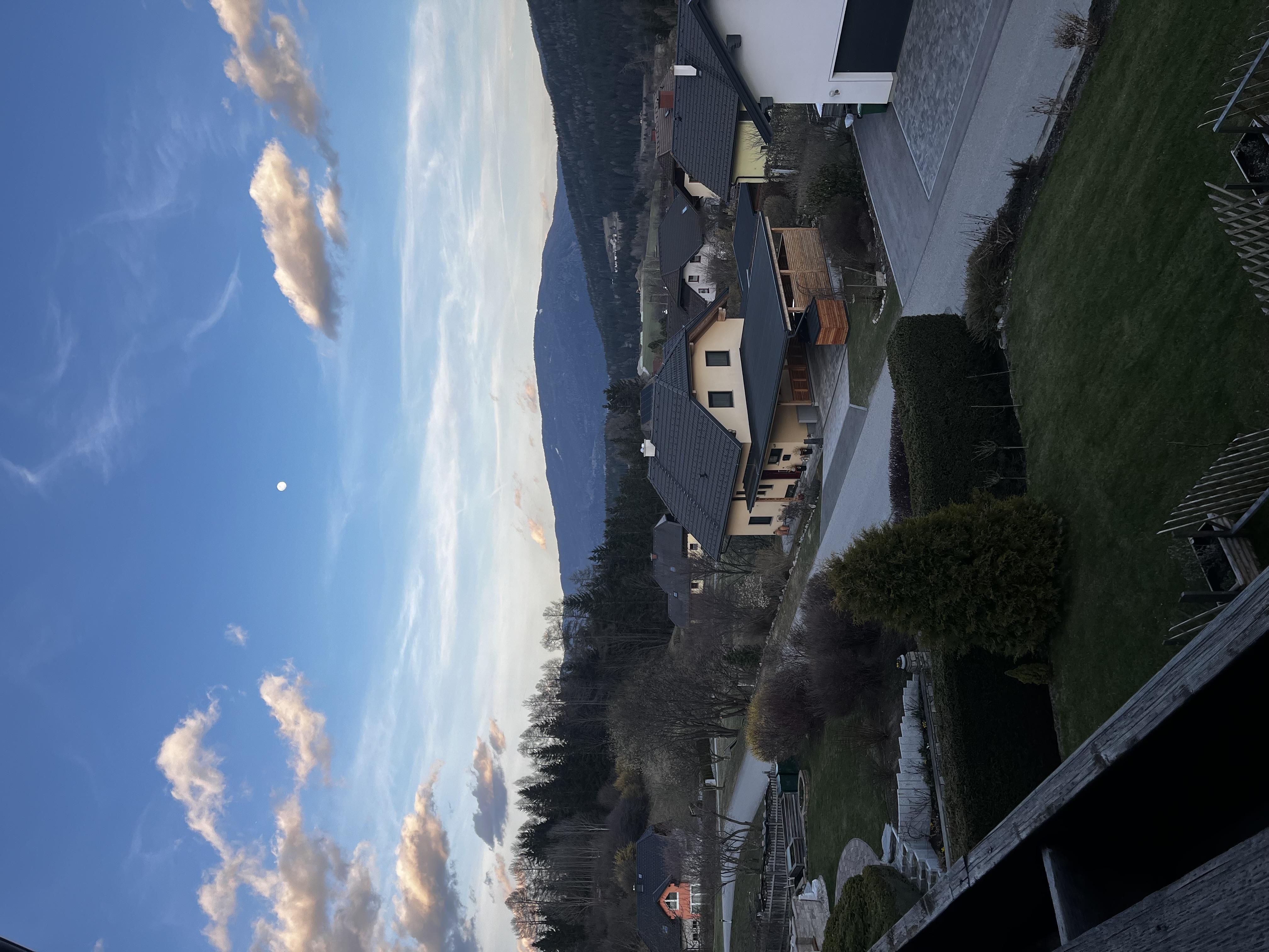 A picturesque view over the mountains with a quiet residential street and houses. The sky is blue with some clouds and the moon is visible.