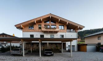 A modern house in alpine style with a large roof and balconies. In front of the building, there are parking spaces and a quiet environment.