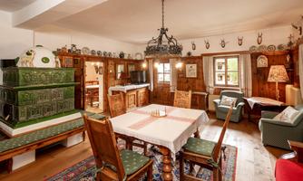 A cozy room with wooden furniture and a large table. The walls are adorned with traditional decorations, and a tile stove stands in the corner.